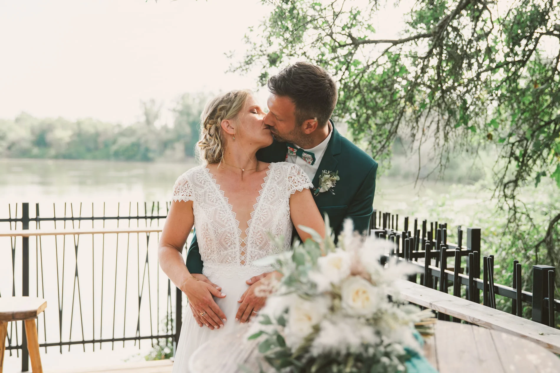 Les mariés s'embrassent sur la terrasse du domaine de Courrèges, à Léguevin. Le bouquet de la mariée est en premier plan et ses couleurs rappellent la robe de la mariée et le costume du marié.