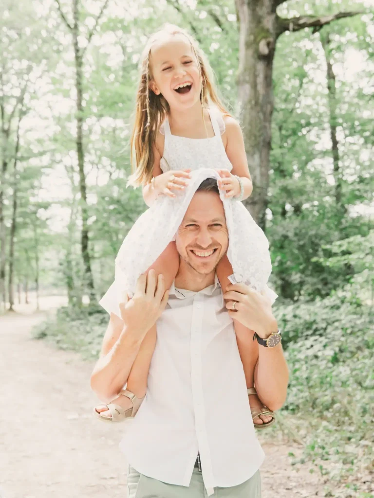 Séance photo famille à Toulouse dans la Forêt de Bouconne. La famille marche dans la forêt. Le père porte sa fille sur ses épaules. La jupe de sa fille remonte sur sa tête ce qui les fait rire aux éclats
