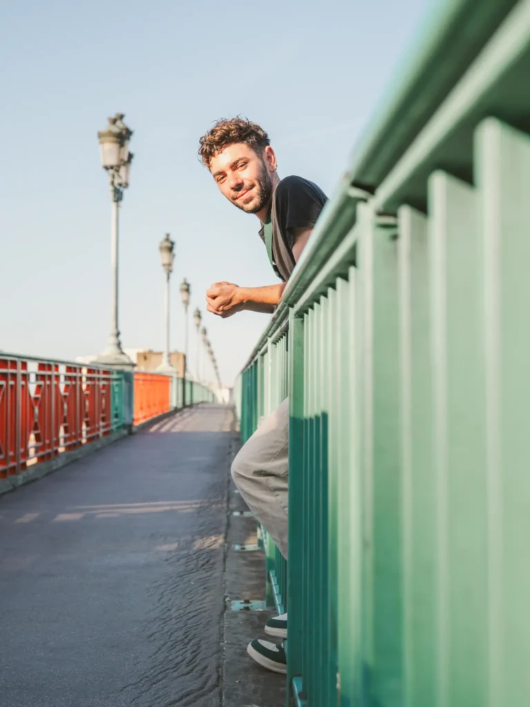 Séance photo portrait à Toulouse sur le pont Saint-Pierre avec Dylan. Accoudé à la rambarde du pont Dylan nous regarde. Le cadrage est tel que seul son pied, son genou ses mains et sa tête sont visibles au travers la rambarde.