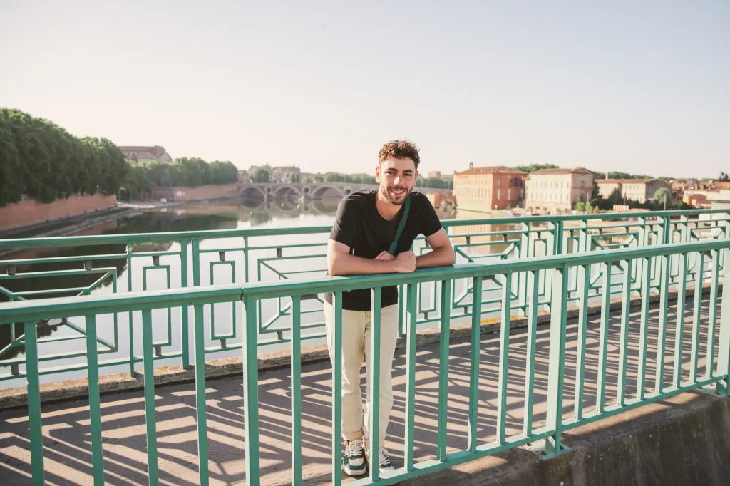 Séance portrait à Toulouse sur le pont Saint-Pierre avec Dylan. Au premier plan, Dylan est accoudé à la barrière et nous regarde en souriant. Derrière lui, la Garonne, le Pont Neuf et les ruines du Pont de la Daurade sont illuminés par les premières lueurs jaunes d'un matin d'été.
