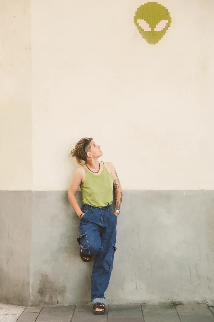Séance portrait à Toulouse sur la place Saint-Pierre avec Marie. Celle-ci est adossée à un mur. Mains dans le dos, un pied contre le mur, elle regarde en hauteur un fine art représentant une tête d'extraterrestre. Cette tête est assortie au haut que porte Marie.