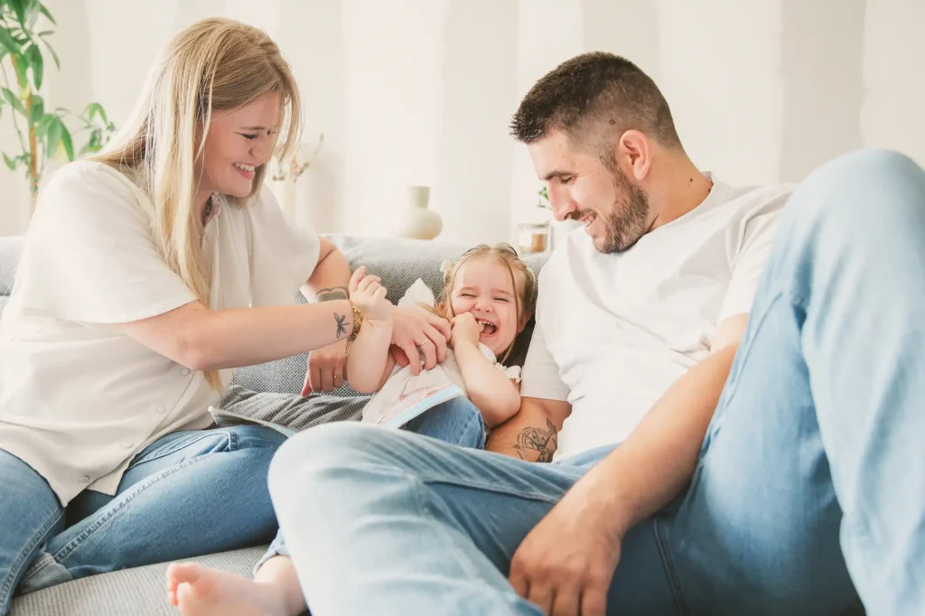Séance photo famille à Toulouse avec des parents et leur fille. Dans leur canapé, ils taquinent leur fille en lui faisant des chatouilles. Celle-ci rie aux éclats ce qui fait rire les parents.