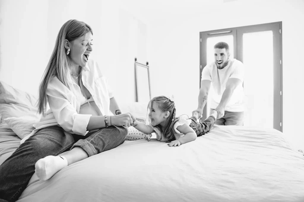 Séance photo grossesse à Toulouse sur le lit des parents. La femme enceinte, assise sur le lit, tend les bras vers sa fille. Celle-ci, allongé de tout son long se fait tirer par les pieds par son père. Des éclats de rire et un moment partagé vient de naître.