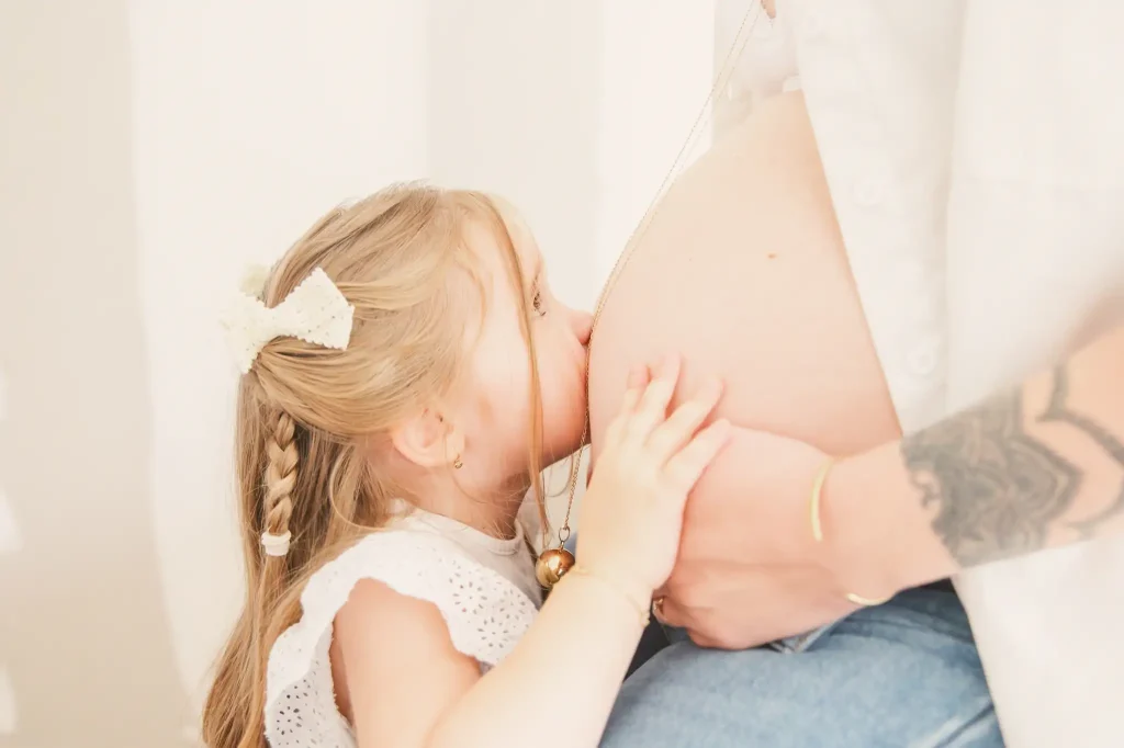 Séance photo grossesse à Toulouse avec la fille de la future maman. La fille de 3 ans fait un bisous sur le ventre bien rebondi de sa mère enceinte.