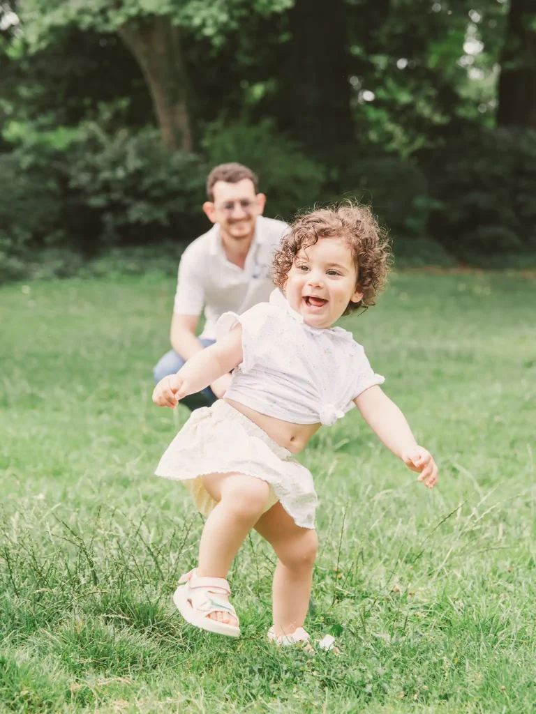 Séance photo famille à Toulouse au jardin des plantes avec une petite fille et ses parents. La fille court partout dans l'herbe du jardin et son père accroupi la regarde se dépenser en souriant.