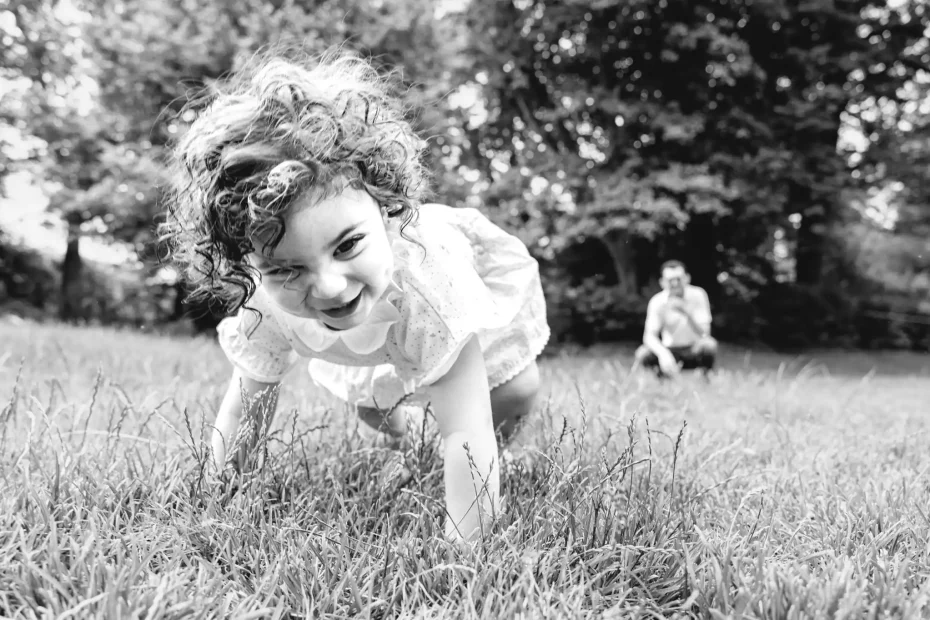 Séance photo famille à Toulouse au jardin des plantes. Le père accroupi dans l'herbe regarde sa fille courir partout, tomber puis se relever en riant au premier plan de la photo.