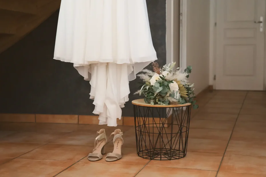 Photographe de mariage à Toulouse au mariage de Franck et Pauline. Le bouquet posé sur une table à coté de la robe de la mariée qui est suspendue au dessus des chaussures.