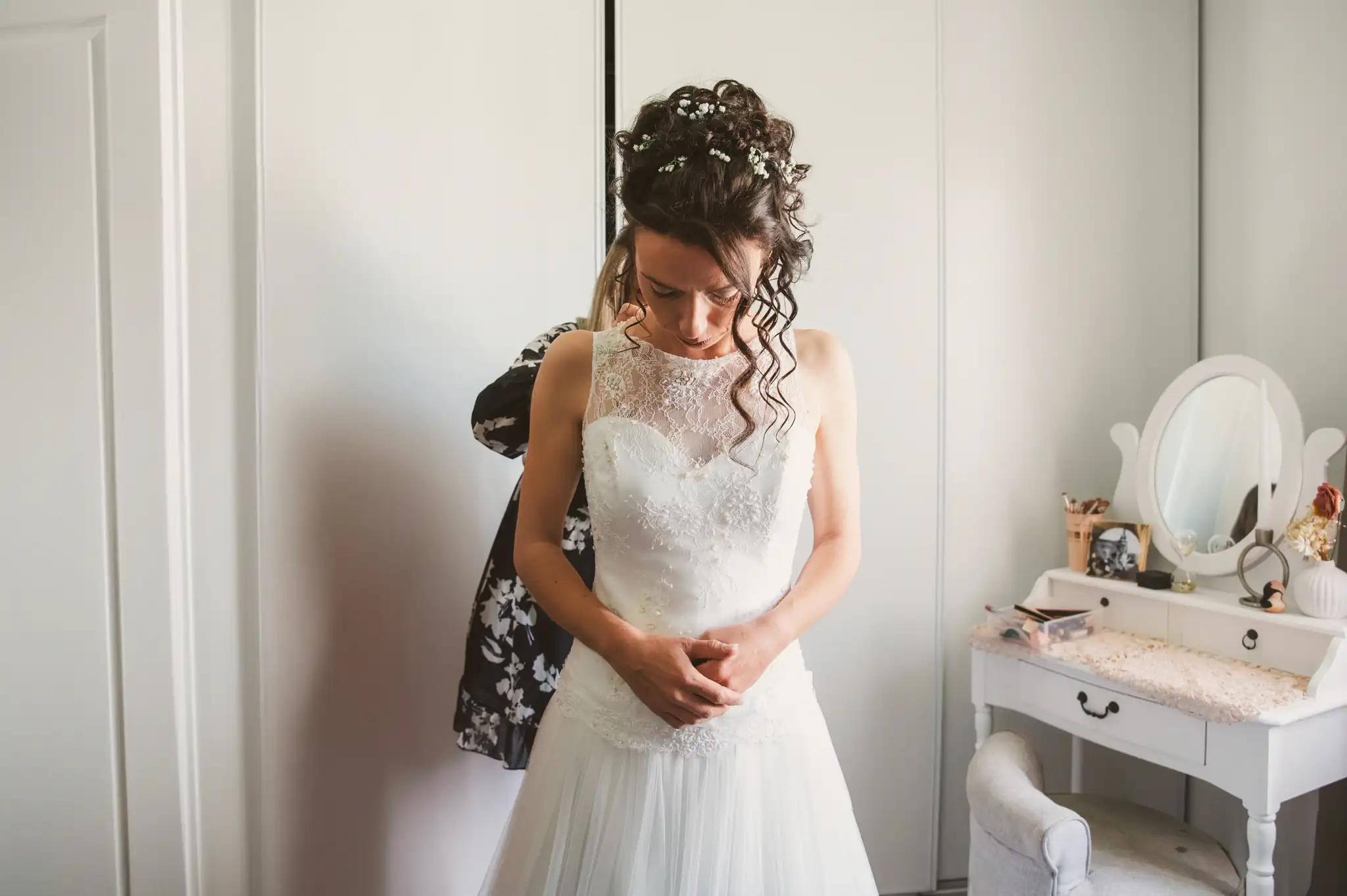 Photographe de mariage à Toulouse lors de l'habillage de la mariée. Une demoiselle d'honneur l'aide à refermer les nombreuses attaches de sa magnifique robe de mariage blanche