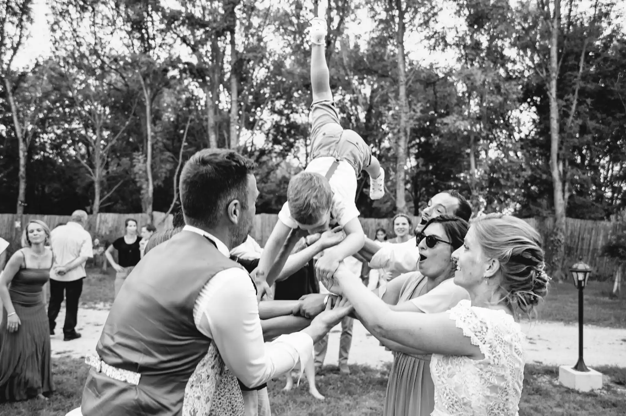 Photographe de mariage à Toulouse au domaine de Courrèges pendant le cocktail. Les invités portent le fils des mariés et le propulsent en l'air pour le faire voler.