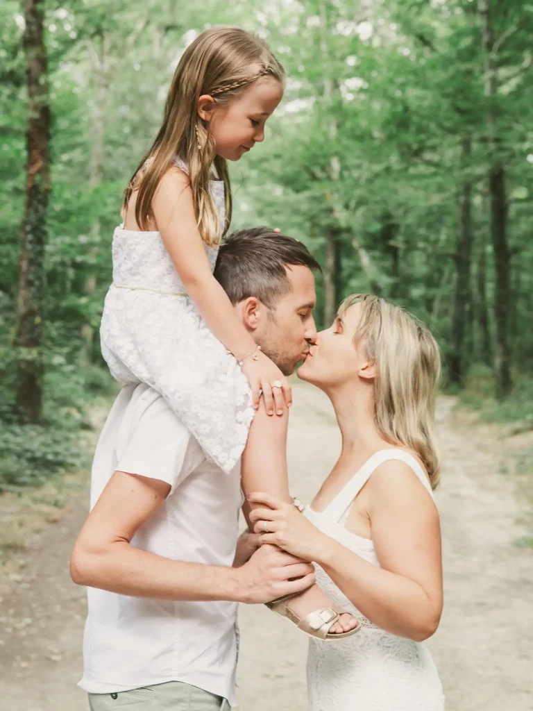 Séance photo famille à Toulouse, à la forêt de Bouconne. Le papa porte sa fille sur les épaules et embrasse sa femme