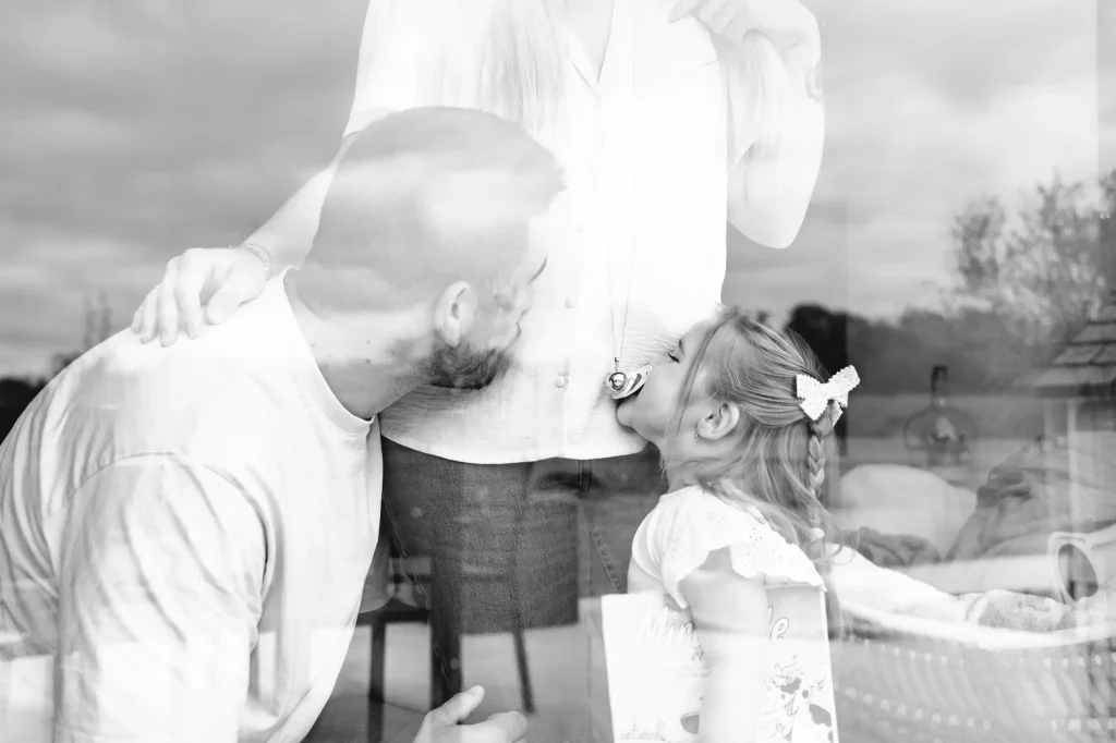 Séance photo grossesse à Toulouse avec le père et sa fille qui embrassent le ventre de la future maman devant la baie vitrée