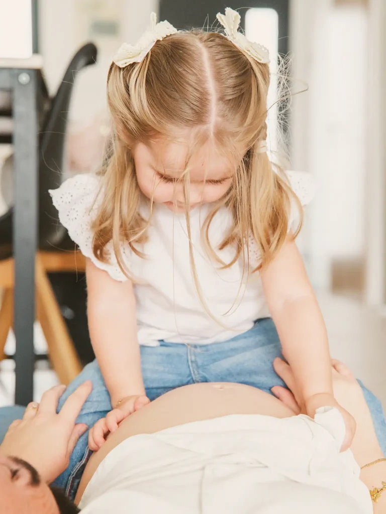 Séance photo grossesse à Toulouse, la fille à cheval sur sa mère regarde son ventre bien arrondi