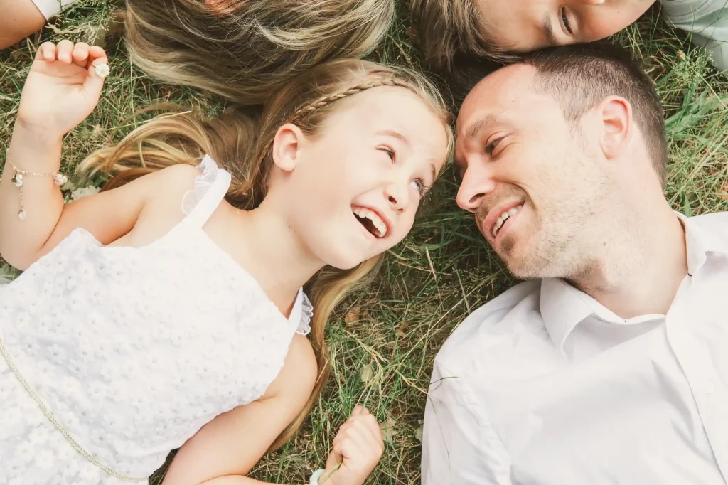 Séance photo famille à Toulouse dans la forêt de Bouconne. Couchés dans l'herbe, le papa et sa fille se regardent en se faisant des clins d’œil.