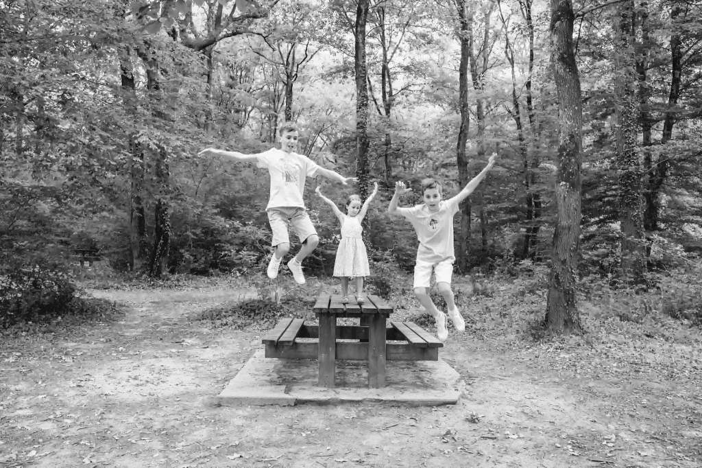 Séance photo famille à Toulouse dans la forêt de Bouconne avec des enfants qui jouent à sauter d'une table de pique-nique en même temps
