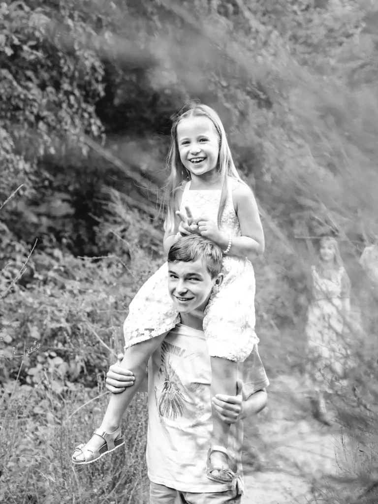 Séance photo famille à Toulouse dans la Forêt de Bouconne avec le grand frère qui porte sa sœur sur les épaules lors d'une balade en bord de lac.