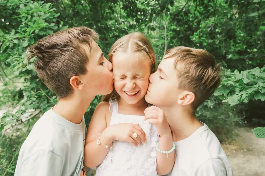 Séance photo famille à Toulouse dans la forêt de Bouconne avec deux frères qui embrassent leur sœur en même temps sur ses joues ce qui la fait rire.