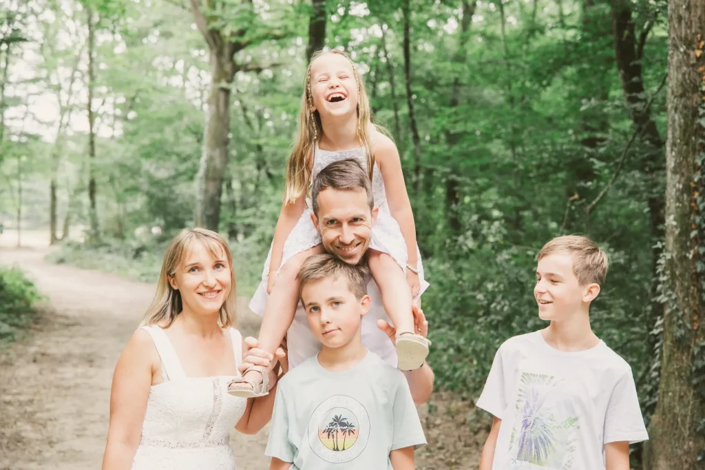 Séance photo famille à Toulouse dans la forêt de Bouconne avec une photo de famille en fin de séance. La fille sur les épaules de son père rit alors que son frère boude.