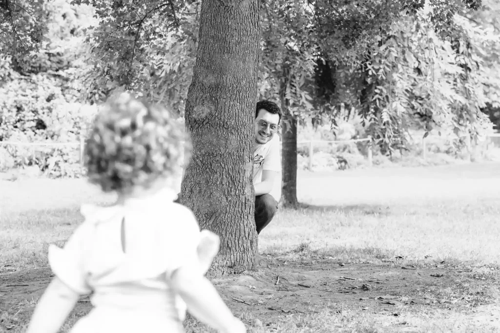 Séance photo famille à Toulouse, au jardin des plantes, avec le papa qui joue à cache-cache avec sa fille derrière un tronc d'arbre.