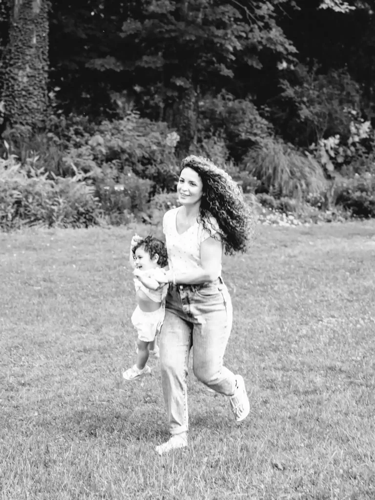 Séance photo famille à Toulouse, au jardin des plantes. La maman attrape sa fille par les bras et court dans l'herbe en la portant.