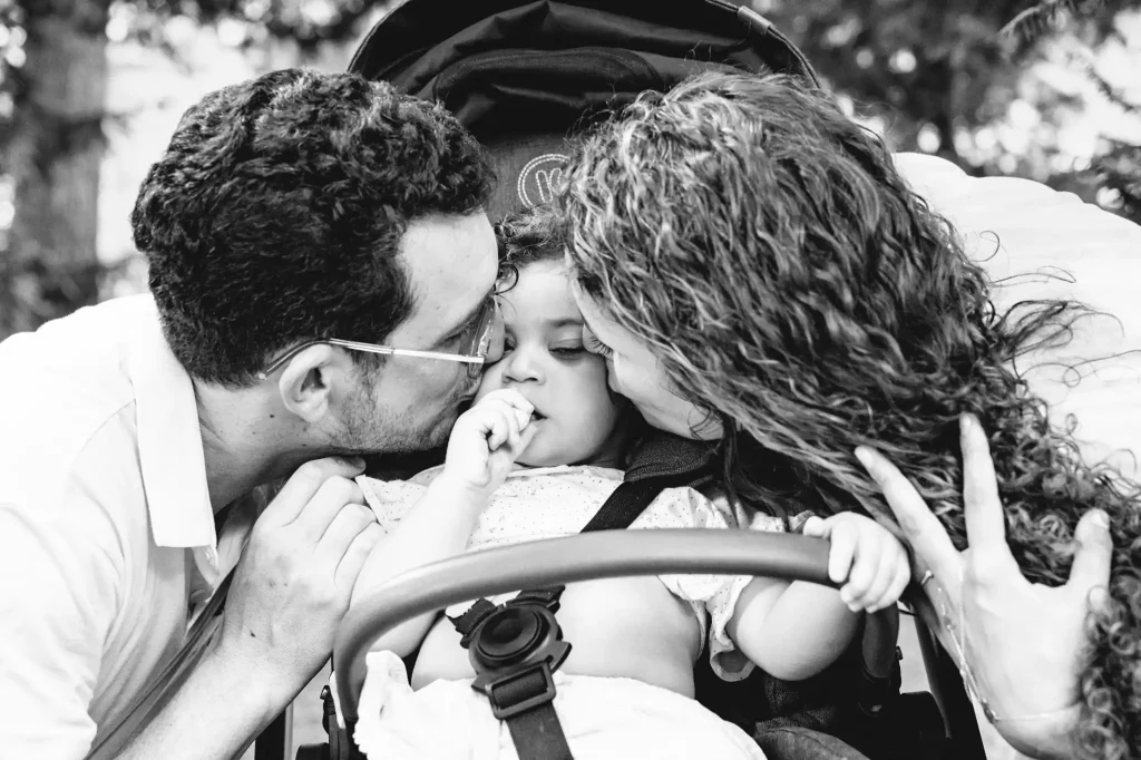 Séance photo famille à Toulouse au jardin des plantes avec les parents qui embrassent leur fille sur chaque joue alors qu'elle mange dans sa poussette.