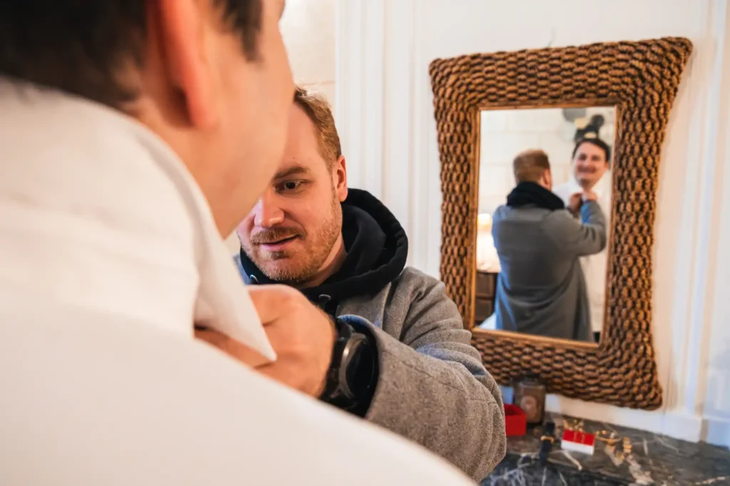 Le témoin aide le marié à fermer sa chemise pendant que celui-ci se regarde dans le miroir en souriant.